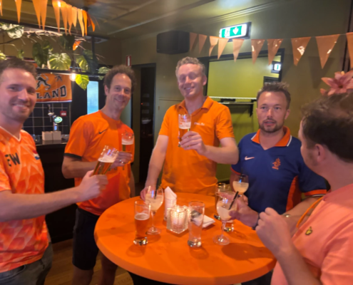 Bezoekers in oranje kleding staan met drankjes rond een tafel tijdens de Oranjeavond.